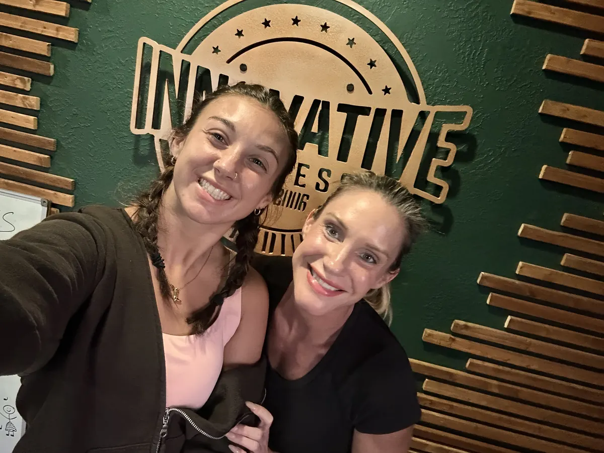 Two women smiling for a selfie in front of the Innovative Fitness logo wall